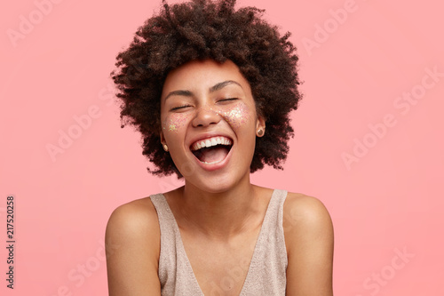Fotografia Joyful African American female with dark skin, laughs happily, opens mouth widely, has sparkles on cheeks, closes eyes, has curly hair, isolated over pink background