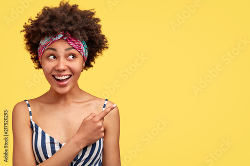 Pretty mixed race female with crisp hair  has gentle smile  shows you something pleasant  indicates with fore finger on blank yellow studio wall. Charming African American woman poses indoor