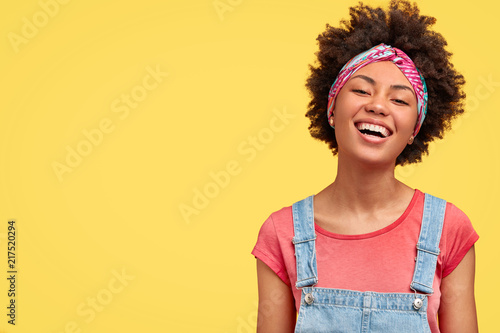 Glad young female with dark skin, white even teeth, laughs positively as sees something funny in front, wears casual t shirt and dungarees, isolated over yellow background with blank space aside