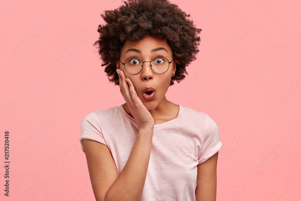 Photo of stupefied dark skinned female touches cheeks with hand, opens eyes widely, dressed in casual t shirt, isolated over pink studio wall. People, surprisement, reaction, emotions concept - obrazy, fototapety, plakaty 