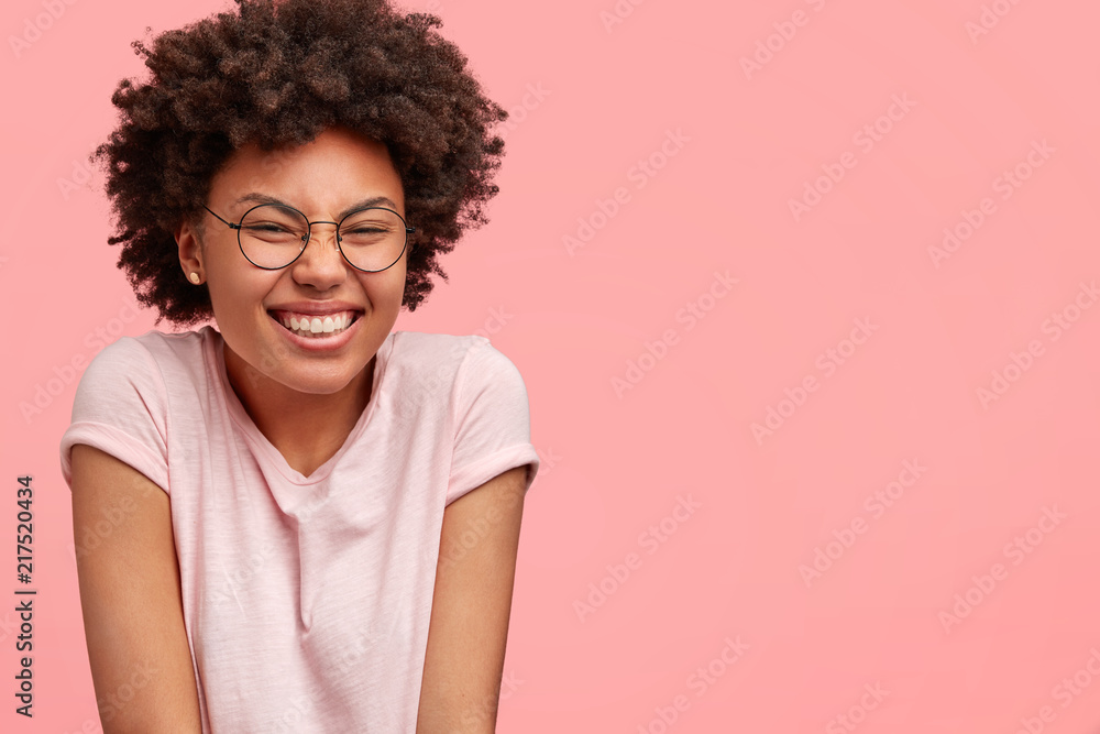 Cheerful dark skinned female student giggles at camera, hears jokes from groupmates, shruggs shoulders, squints face, dressed casually, models in studio against pink background with free space Cheerful dark skinned female student giggles at camera, hears jokes from groupmates, shruggs shoulders, squints face, dressed casually, models in studio against pink background with free space