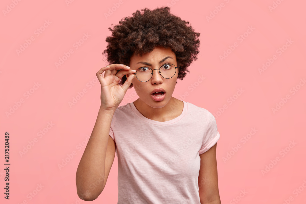 Horizontal shot of attractive African American female with scrupulous expression, looks with indignation through eyewear, opens mouth, tries to read something from distance, poses indoor alone Horizontal shot of attractive African American female with scrupulous expression, looks with indignation through eyewear, opens mouth, tries to read something from distance, poses indoor alone
