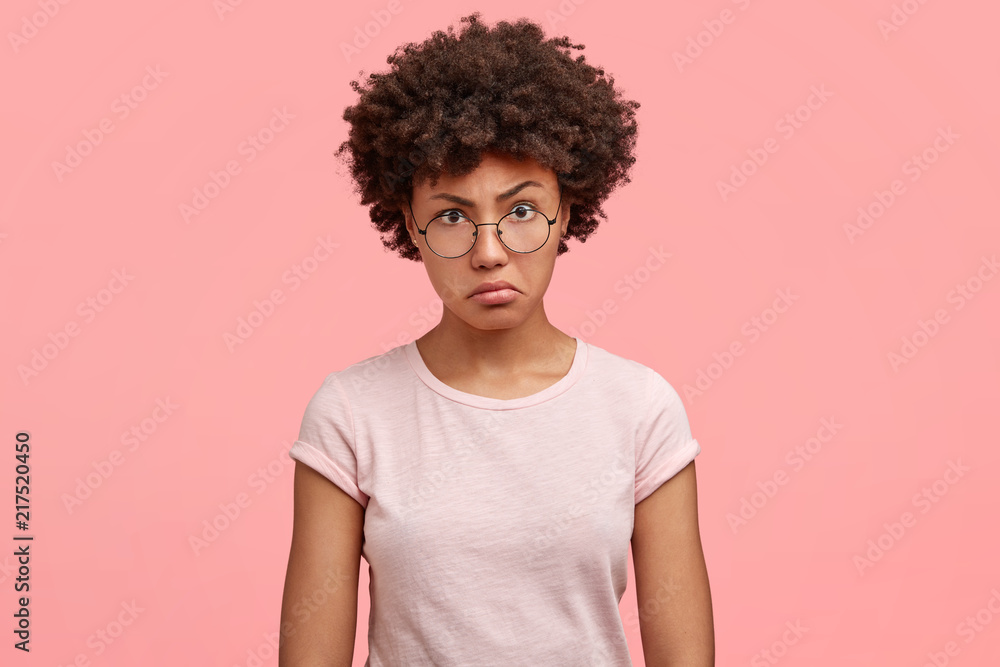 Waist up portrait of dissatisfied African American female with negative angry facial expression, looks in displeasure at someone, wears casual t shirt, being annoyed spend weekend at home alone Waist up portrait of dissatisfied African American female with negative angry facial expression, looks in displeasure at someone, wears casual t shirt, being annoyed spend weekend at home alone
