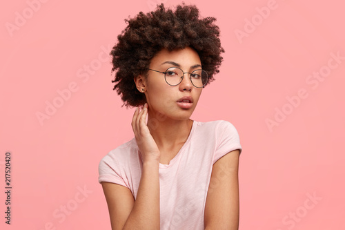 Sideways shot of adorable young female with dark skin, looks seriously and confidently at camera, shows her natural beauty, poses for fashion magazine, wears round glasses and casual t shirt