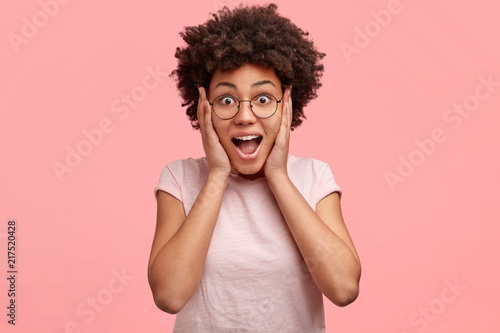 Indoor shot of surprised glad female with appealing appearance, black curly hair and dark skin, stares with eyes full of disbelief, keeps hands on cheeks, recieves something unexpected and expensive