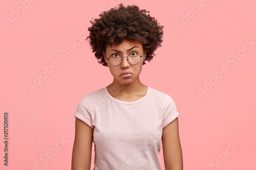 Waist up portrait of dissatisfied African American female with negative angry facial expression, looks in displeasure at someone, wears casual t shirt, being annoyed spend weekend at home alone