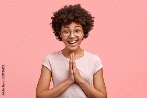 Image of happy African American female with pleasant smile, dressed casually, presses hands together, poses in praying gesture, beggs for forgiveness with joyful expression, isolated on pink wall