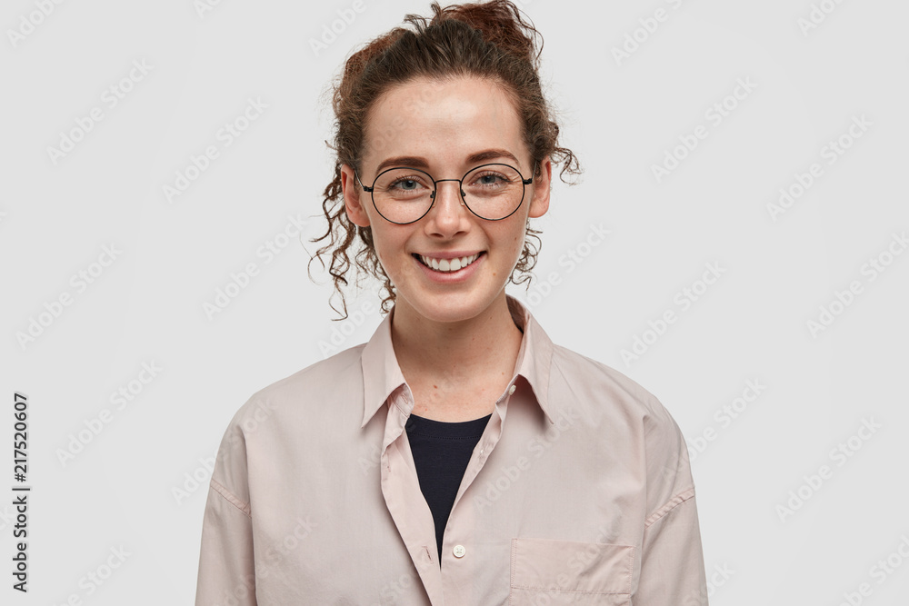 Studio shot of positive European young female with dark curly hair, has gentle smile, freckled skin, wears casual beige shirt, models against white background, being glad to be promoted at work Studio shot of positive European young female with dark curly hair, has gentle smile, freckled skin, wears casual beige shirt, models against white background, being glad to be promoted at work