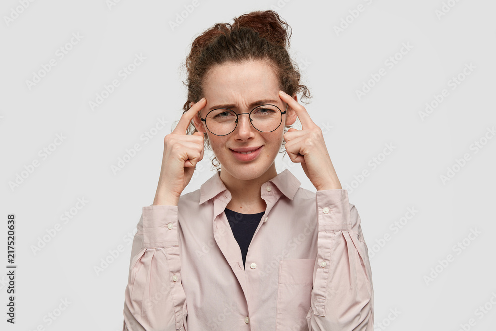 Stressful young female frowns face, keeps fore fingers on temples, tries to conetrate on something, has round eyewear and beige shirt, isolated over white background. Frustrated concerned woman Stressful young female frowns face, keeps fore fingers on temples, tries to conetrate on something, has round eyewear and beige shirt, isolated over white background. Frustrated concerned woman