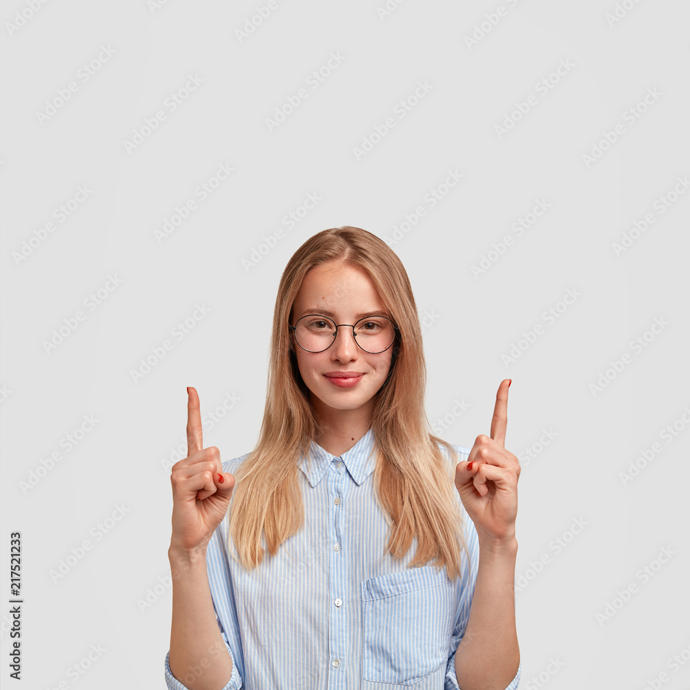 Vertical shot of satisfied young European female with pleased expression, points upwards with index fingers, dressed in fashionable shirt, shows something above, isolated over white background. Vertical shot of satisfied young European female with pleased expression, points upwards with index fingers, dressed in fashionable shirt, shows something above, isolated over white background.