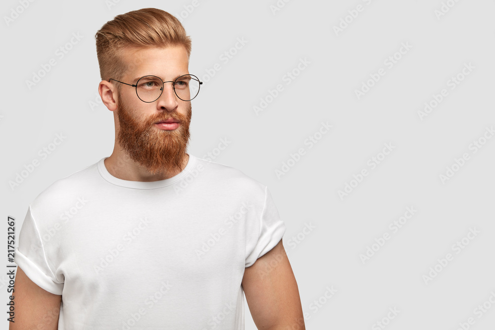 Brutal young handsome man with ginger beard, trendy hairstyle, wears casual white t shirt and spectacles, looks aside, stands against white background with free space for your advertisement. Brutal young handsome man with ginger beard, trendy hairstyle, wears casual white t shirt and spectacles, looks aside, stands against white background with free space for your advertisement.