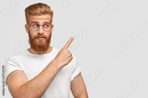Horizontal shot of attractive Caucasian male has ginger hair and beard, points with fore finger aside, dressed in t shirt in one colour with background, shows blank space for your advertisement