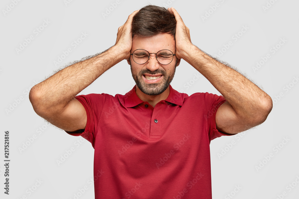 Fototapeta premium Horizontal shot of fatigue young Euroepean unshaven male keeps hands on head, feels exhausted after work in office, has headache, wears casual bright red t shirt, isolated over white background