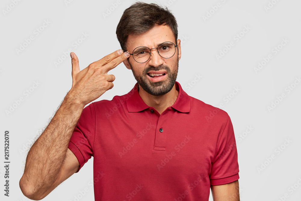 Fototapeta premium Young stressful male makes gun gesture, pretends commiting suicide, keeps two fingers on temples, feels desperate, poses alone against white background, dressed in casual red t shirt, has crisis