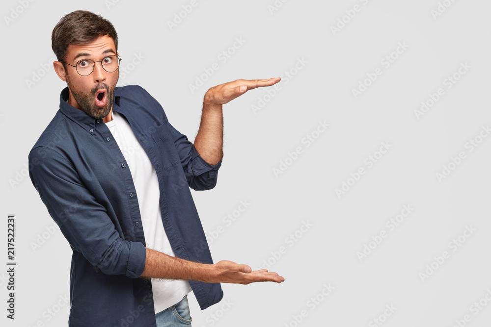 Stunned unshaven male with shocked facial expression gestures with hands, shows size or height of something, dressed in fashionable shirt, isolated over white background, copy space for your text - obrazy, fototapety, plakaty 