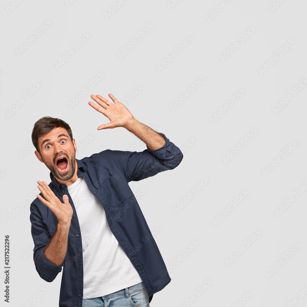 Funny surprised amazed male opens mouth widely, gestures with hands, notices something wonderful, wears in casual t shirt and dark blue shirt, poses against white wall with blank space for your text Funny surprised amazed male opens mouth widely, gestures with hands, notices something wonderful, wears in casual t shirt and dark blue shirt, poses against white wall with blank space for your text