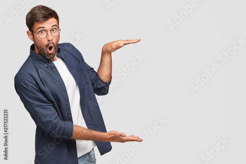 Stunned unshaven male with shocked facial expression gestures with hands, shows size or height of something, dressed in fashionable shirt, isolated over white background, copy space for your text