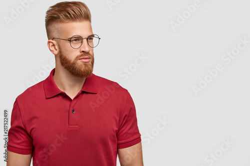 Sideways shot of ginger male with trendy haircut, thick beard, focused aside, thinks about new project, wears bright casual red t shirt, isolated over white background with copy space for your text © wayhome.studio 