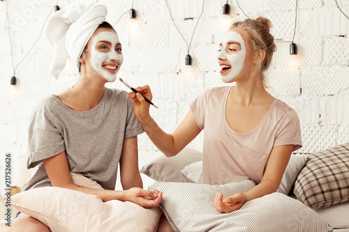 Portrait of attractive happy girls having fun sitting in bed with pillows. Cheerful women wearing facial cosmetic mask for soft smooth silk skin. Beauty skincare concept