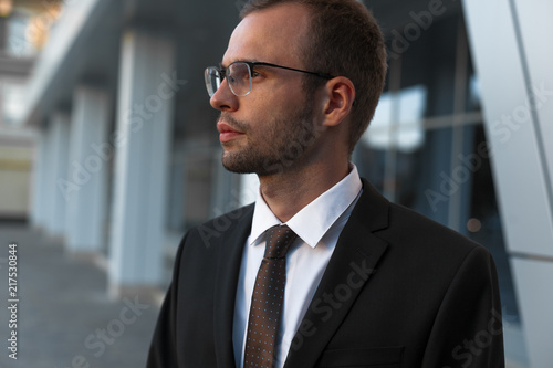 Young businessman in official black suit is standing in front of office building