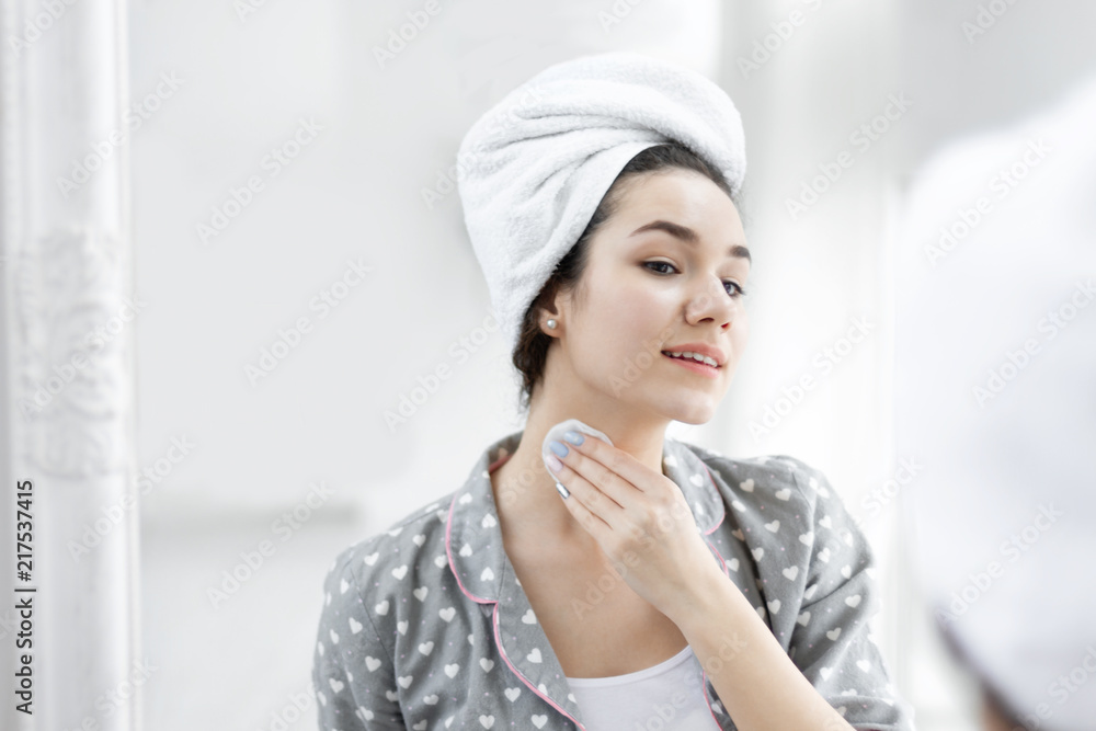 Pretty young brunette woman female girl after shower wearing pajamas sleepwear and white towel over her head washing out make up with cotton pads in front of mirror. Morning / body skin care concept Pretty young brunette woman female girl after shower wearing pajamas sleepwear and white towel over her head washing out make up with cotton pads in front of mirror. Morning / body skin care concept