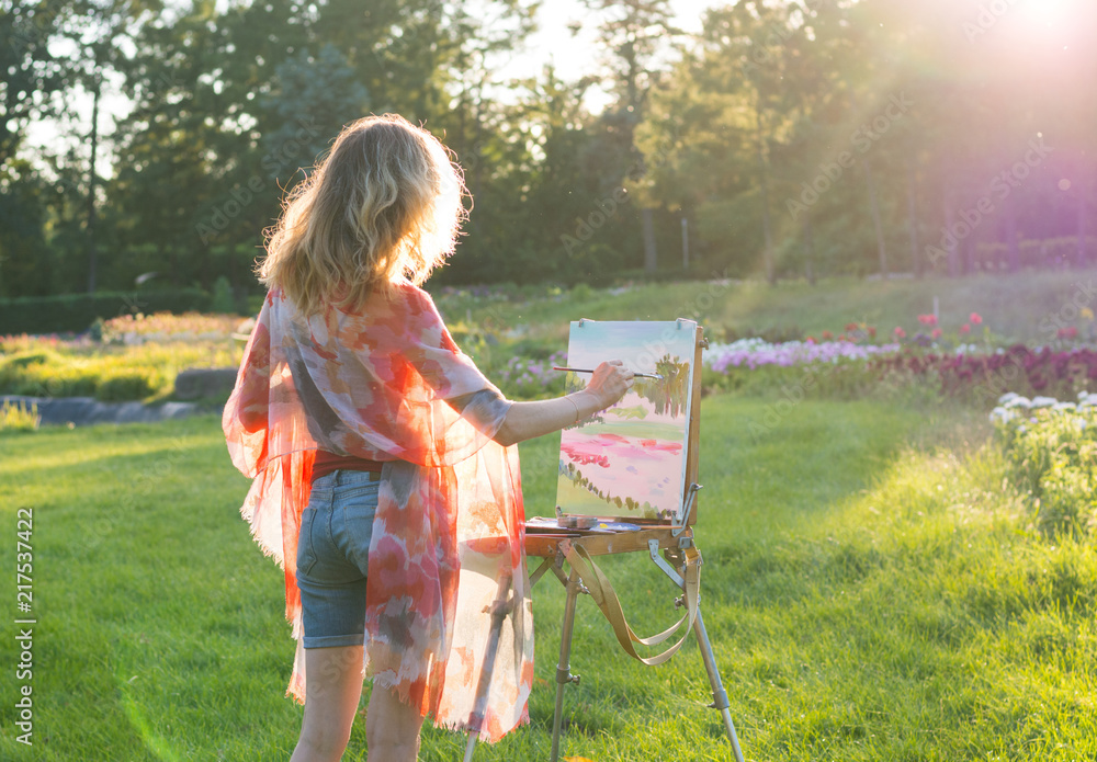 A young woman artist holds a brush and paints a picture on an easel in the rays of the sunset.