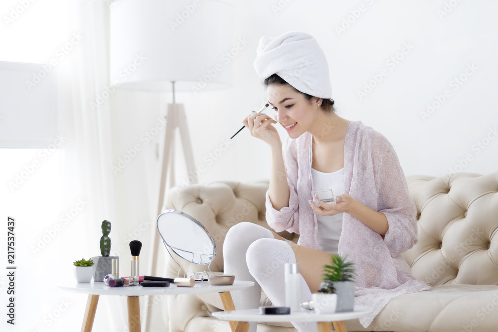 Young girl female woman after shower wearing white underwear and white towel sitting on sofa and doing make up / painting brows in front of mirror in light interior room. Preparing for new day concept Young girl female woman after shower wearing white underwear and white towel sitting on sofa and doing make up / painting brows in front of mirror in light interior room. Preparing for new day concept
