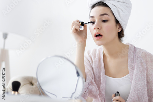 Young girl female woman after shower wearing white underwear and white towel sitting on sofa and doing make up / painting eyelashes in front of mirror in light room. Preparing for new day concept