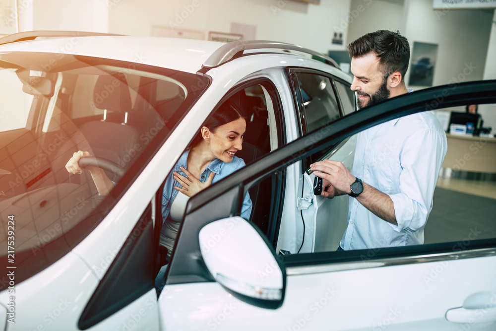 Beautiful young modern couple chooses and buying a new car for the family in the dealership. Woman inside of car tests it Beautiful young modern couple chooses and buying a new car for the family in the dealership. Woman inside of car tests it