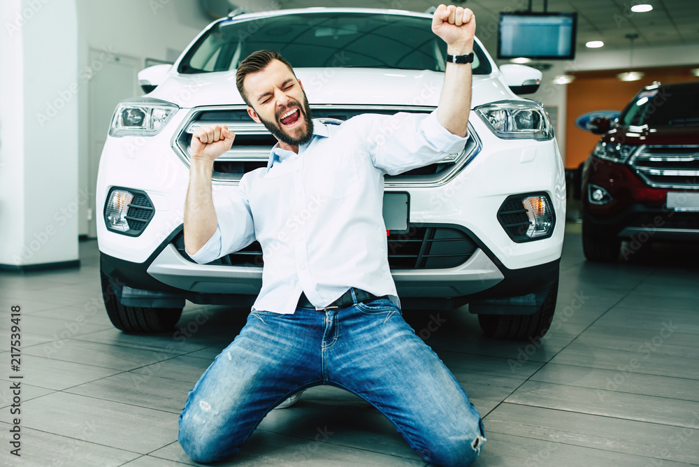 Happy handsome man buying a new white car in dealership and dancing on the floor against the auto. - obrazy, fototapety, plakaty 