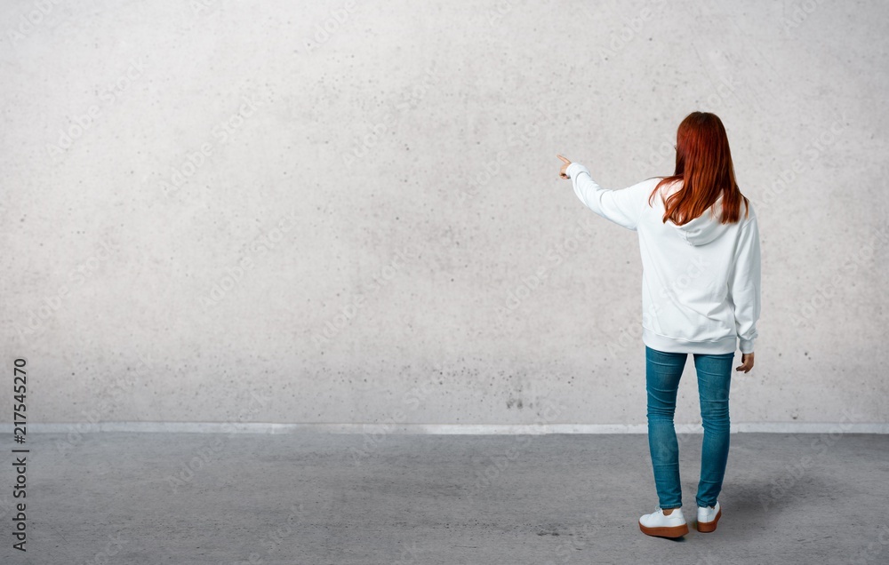 Young redhead girl in an urban white sweatshirt with glasses pointing back with the index finger presenting a product from behind and standing on a vintage gray wall - obrazy, fototapety, plakaty 