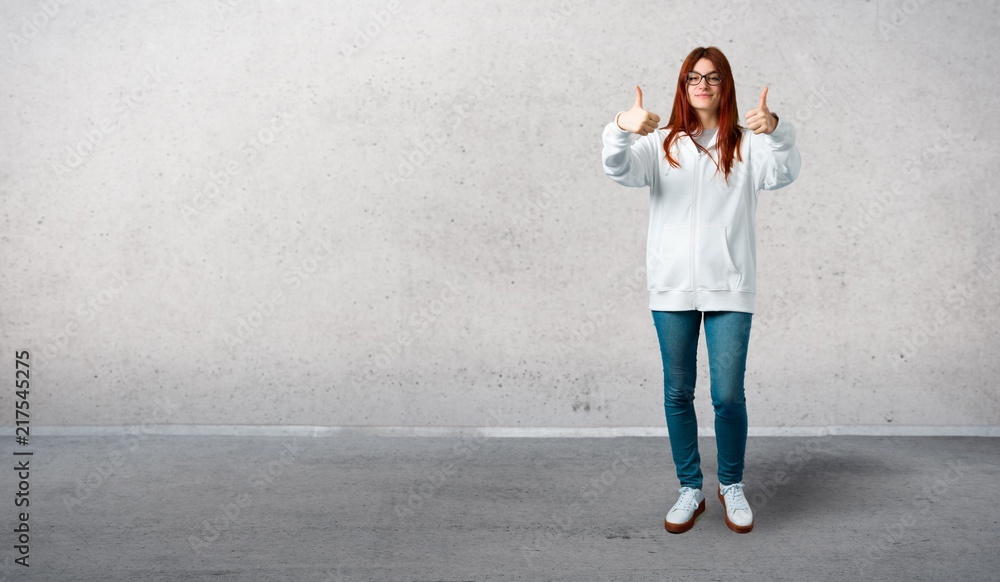 Young redhead girl in an urban white sweatshirt with glasses giving a thumbs up gesture with both hands and smiling. Cheerful expression and standing on a vintage gray wall Young redhead girl in an urban white sweatshirt with glasses giving a thumbs up gesture with both hands and smiling. Cheerful expression and standing on a vintage gray wall