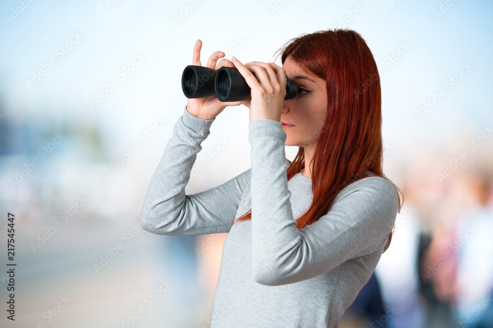 Young redhead girl and looking for something in the distance with binoculars on unfocused background