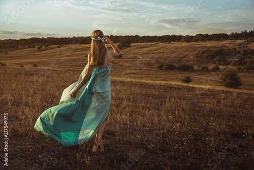 A girl in a long dress is walking along the field at sunset