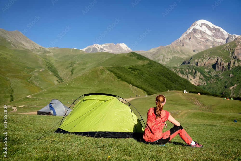 Fototapeta premium Treking for the Kazbek peak in the mountains of the Caucasus. the tourist is sitting on the grass by the tent and is looking at the snowy peak.