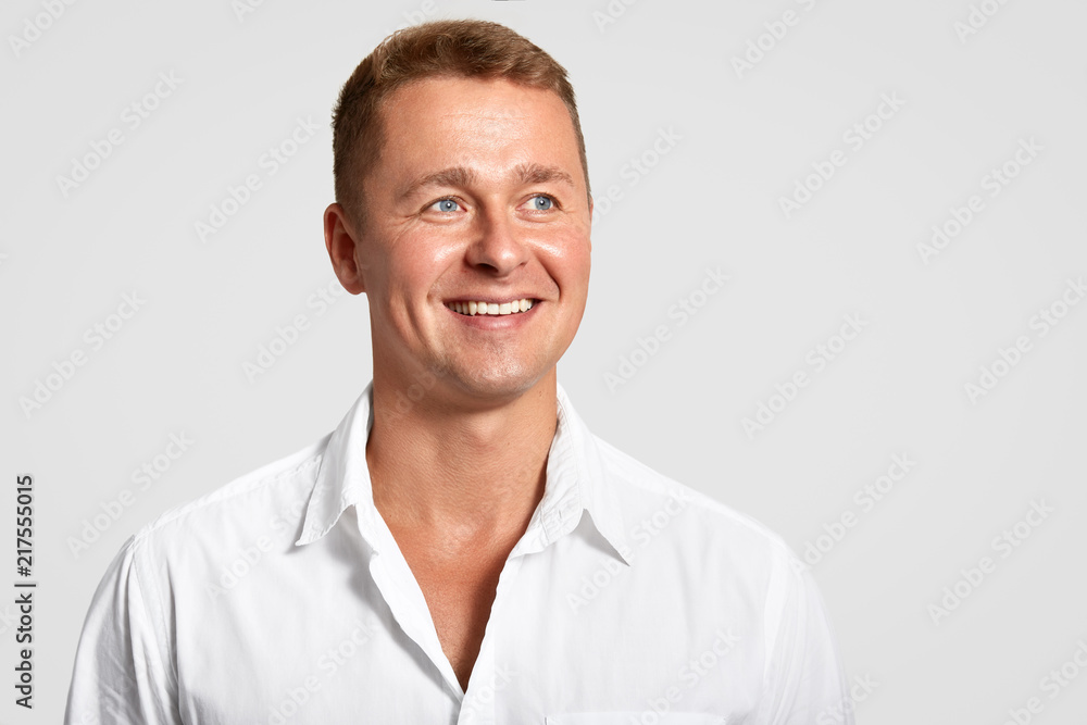 Dremy blue eyed male with positive expression, broad smile, shows white teeth, looks into distance, thinks about something, wears elegant shirt, isolated over white background with blank space