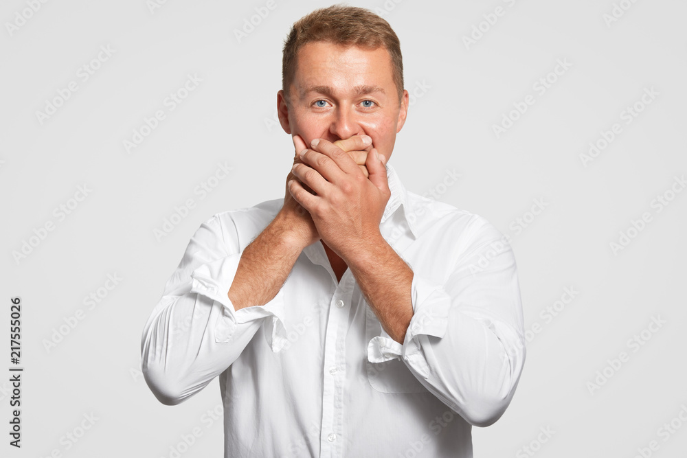 Handsome young male with blue eyes and healthy skin, covers mouth with both palms, tries to be speechless, doesnt want to reveal secret, dressed in elegant white shirt in one tone with background Handsome young male with blue eyes and healthy skin, covers mouth with both palms, tries to be speechless, doesnt want to reveal secret, dressed in elegant white shirt in one tone with background