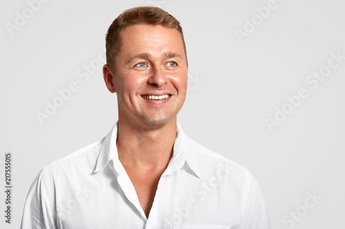 Dremy blue eyed male with positive expression, broad smile, shows white teeth, looks into distance, thinks about something, wears elegant shirt, isolated over white background with blank space