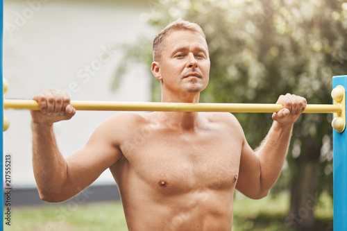 Outside shot of determined young sportsman with muscular body, trains on horizontal bar, has thoughtful expression, dreams about nice biceps and stong health. People and active lifestyle concept