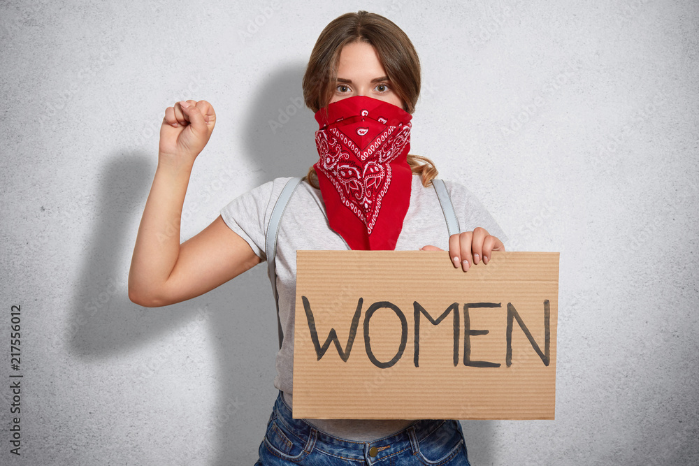 Feminism concept. Self confident young woman feminist protects womens rights, takes part in protest, keeps hands in fist raised, wears red bandana and grey t shirt, stands being serious Stock Photo