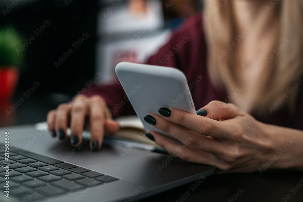 Selective focus on hand holding smartphone in white silicone case. Young female reading text message on mobile phone device. Woman freelance synchronizing her cell phone with laptop computer. Selective focus on hand holding smartphone in white silicone case. Young female reading text message on mobile phone device. Woman freelance synchronizing her cell phone with laptop computer.