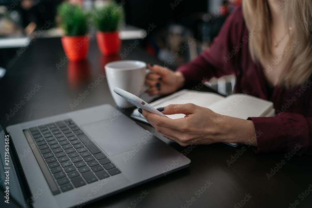 Close up view of female person hands holding mobile telephone while sitting at table with cup of morning coffee and laptop computer. Cropped view of woman reading news via application on smartphone