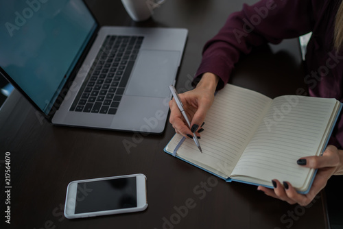 Female student takes notes and writes them to notebook while searching useful information in Internet. Selective focus on woman's hand with pen records planned schedule in notepad.