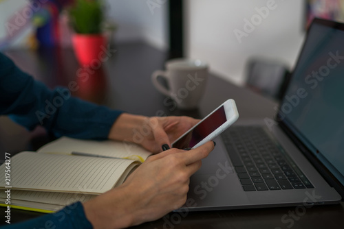Selective focus on modern smartphone device in women's hands. Cropped view of female person doing multimedia data transfer while connecting devices, as mobile phone and laptop computer.