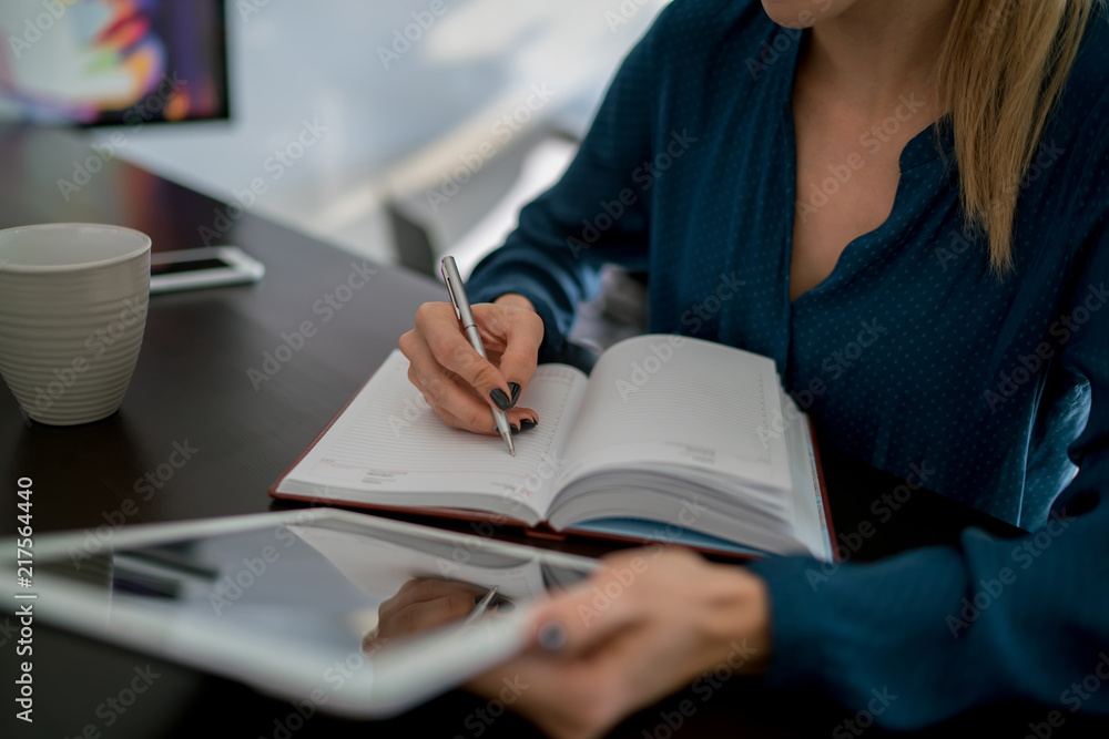 Close up view with selective focus on woman's hand with silver pen over planning schedule notebook. Female student learning online while reading literature from website on digital tablet Close up view with selective focus on woman's hand with silver pen over planning schedule notebook. Female student learning online while reading literature from website on digital tablet