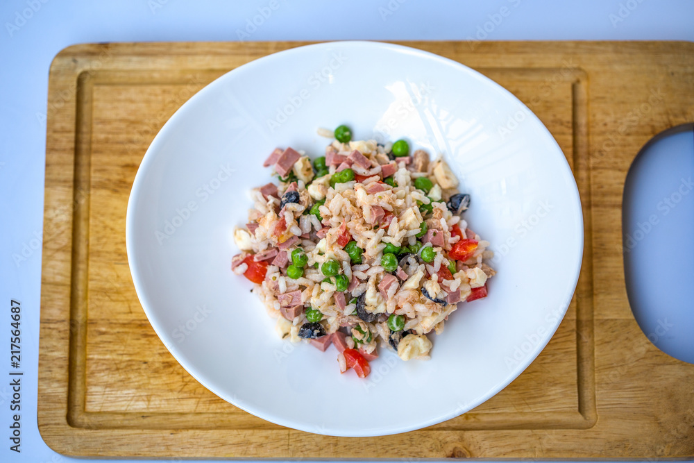 Italian rice salad or cold rice in a white plate on a chopping board with blurred white background. Italian rice salad or cold rice in a white plate on a chopping board with blurred white background.