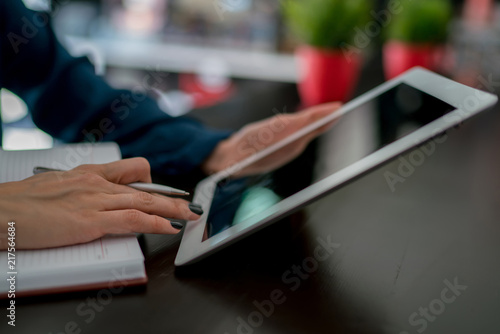 Cropped view of businesswoman writes information in notepad while using tablet with her finger pressed on the button. Female blogger working on new theme for article making text blanks in a notebook