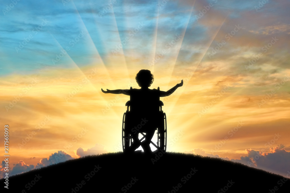 Silhouette of a happy disabled child girl sitting in a wheelchair atop a hill at sunset