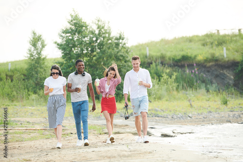 Group of friends enjoying leisure time on fresh air outdoors. They walking with drinks in non-urban area