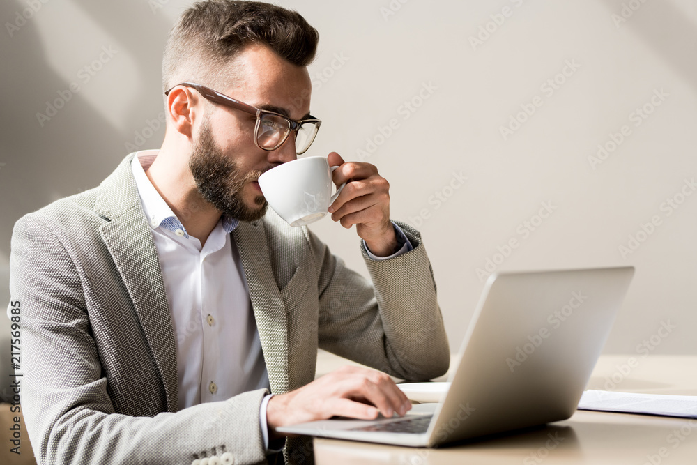 Young bearded businessman in stylish suit sitting at the desk and working on laptop while drinking coffee Young bearded businessman in stylish suit sitting at the desk and working on laptop while drinking coffee
